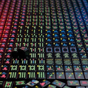 May include: A collection of black Mahjong tiles arranged in a grid, featuring vibrant neon-colored engravings of Chinese characters, numbers, directional symbols, and floral patterns, with some tiles scattered at the bottom of the frame.