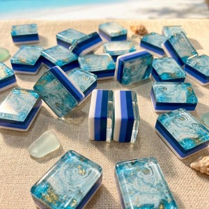 May include: A collection of rectangular mahjong tiles featuring a marbled blue and white resin top with embedded gold flakes, layered over solid blue and white base sections. The tiles are arranged in a scattered pattern on a textured surface.