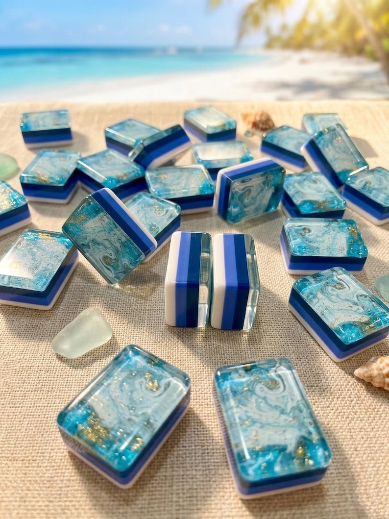 May include: A collection of rectangular mahjong tiles featuring a marbled blue and white resin top with embedded gold flakes, layered over solid blue and white base sections. The tiles are arranged in a scattered pattern on a textured surface.