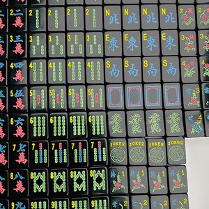 May include: A collection of black mahjong tiles arranged in a grid. The tiles feature colorful engraved characters, numbers, and symbols including JOKER, N, E, S, and various floral and dragon motifs in shades of blue, green, red, and yellow.