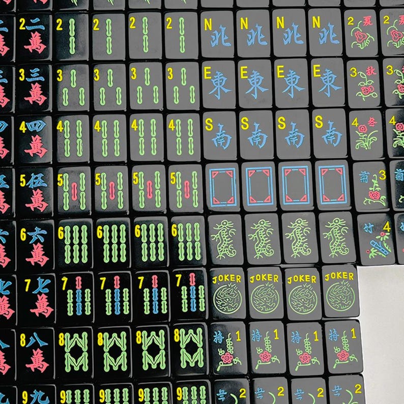 May include: A collection of black mahjong tiles arranged in a grid. The tiles feature colorful engraved characters, numbers, and symbols including JOKER, N, E, S, and various floral and dragon motifs in shades of blue, green, red, and yellow.