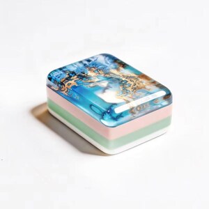 May include: A rectangular resin paperweight featuring a top layer with a swirling blue, white, and gold metallic pattern. The base consists of three distinct horizontal layers in solid pastel pink, light green, and white, creating a smooth finish.
