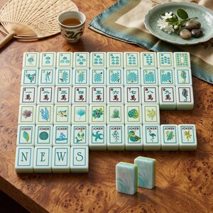May include: A full Mahjong set with white tiles displaying diverse symbols and Chinese characters. The tiles are arranged on a wooden surface, with a cup of tea, a fan, and decorative elements in the background. The word "NEWS" is spelled out with tiles.