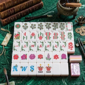 Puede incluir: Un juego de fichas de Mahjong blancas dispuestas en cuadr&iacute;cula, con grabados coloridos de flores, bamb&uacute;, dragones, n&uacute;meros chinos y s&iacute;mbolos de Joker. Las fichas se muestran sobre una superficie verde junto a libros e incienso.