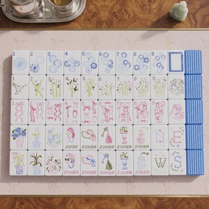 May include: A set of white mahjong tiles arranged in a grid, featuring blue, green, and pink illustrations of flowers, ribbons, birds, hats, and keys. The tiles include numbered suits, joker tiles, and directional markers labeled N, E, W, S.