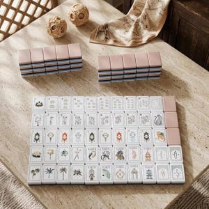 May include: A mahjong set featuring white tiles with various illustrations including numbers, symbols, and animals, alongside two stacks of pink-backed tiles, two round textured spheres, and a beige fabric piece with a printed map design.