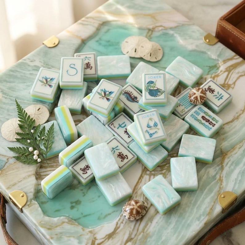 May include: A selection of light blue and white Mahjong tiles, featuring diverse symbols and illustrations, such as flowers and the word "Joker." The tiles are displayed on a decorative, marbled surface, accompanied by small seashells and a fern.