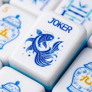 Op de afbeelding: Een witte rechthoekige mahjongtegel met een blauwe afbeelding van een vis en het woord JOKER in blauwe letters. De tegel wordt omringd door andere mahjongtegels, waaronder enkele met blauwe en gele illustraties van vazen en Chinese karakters.