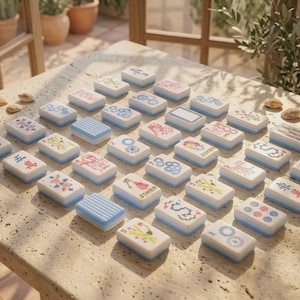 May include: A collection of rectangular mahjong tiles with white faces and light blue sides, featuring various colorful illustrations, symbols, and the word JOKER. The tiles are arranged in a grid pattern on a textured stone surface.