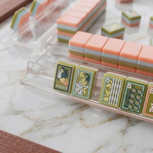 May include: A set of rectangular mahjong tiles with pastel peach, light blue, and olive green layers, arranged in clear plastic racks. The tiles feature detailed illustrations including a joker, a peacock, and the number eight in olive green.