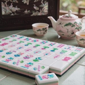 May include: A set of white mahjong tiles with colorful floral and character engravings, including tiles labeled JOKER. The tiles feature a layered design with white, light green, and pink sections, arranged in a grid with one tile placed in front.