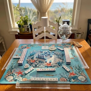 May include: A mahjong set featuring white tiles with blue and multi-colored illustrations, arranged on a light blue play mat decorated with coral reef, mermaid, and sea life imagery. Includes four clear acrylic tile racks and a small white lamp.