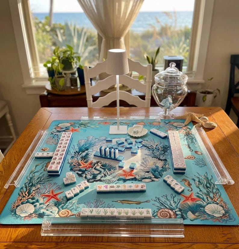 May include: A mahjong set featuring white tiles with blue and multi-colored illustrations, arranged on a light blue play mat decorated with coral reef, mermaid, and sea life imagery. Includes four clear acrylic tile racks and a small white lamp.