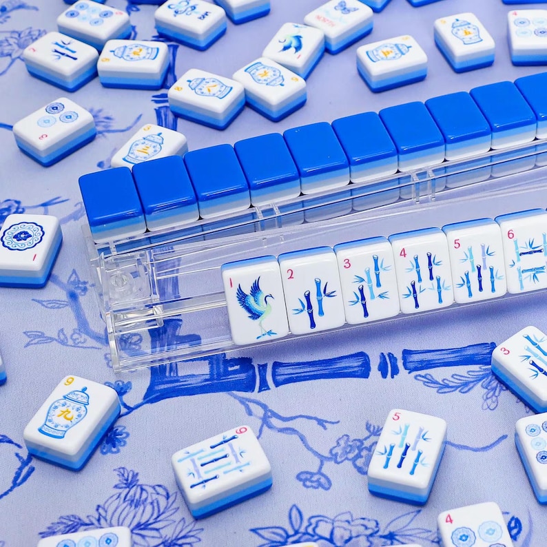 Op de afbeelding: Een set rechthoekige Mahjong-tegels met blauw-witte ontwerpen, waaronder afbeeldingen van vogels, bamboestengels en vazen. Sommige tegels zijn gerangschikt in een doorzichtige acrylhouder, terwijl andere verspreid liggen op een blauw bloemenoppervlak.