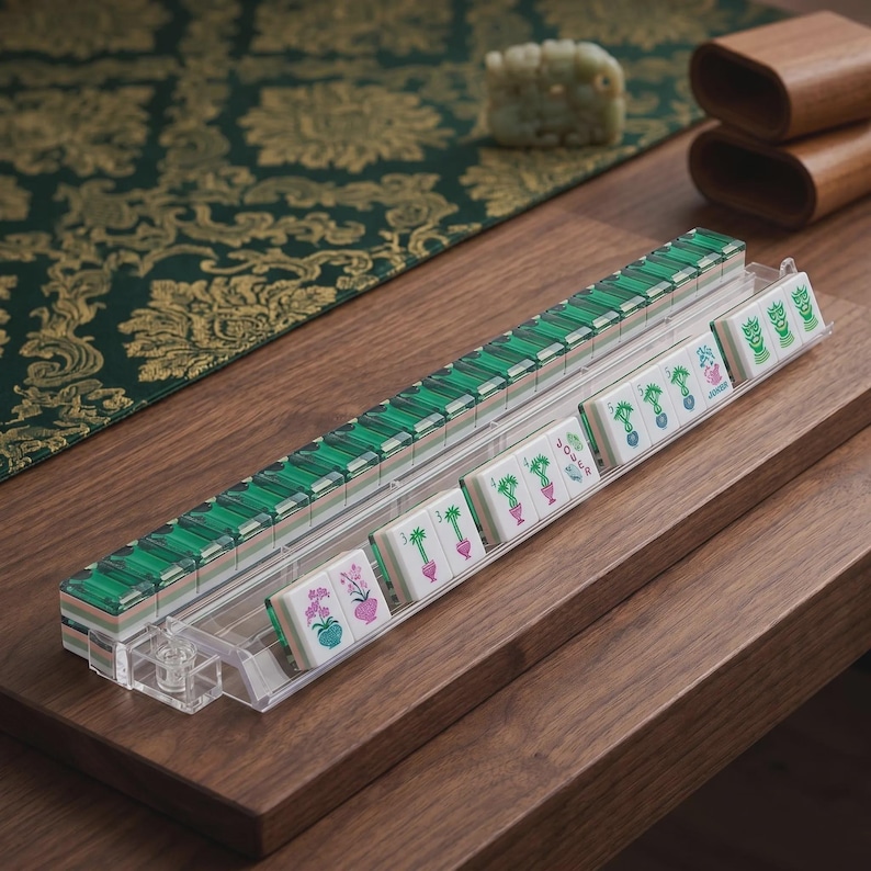 May include: A clear acrylic Mahjong tile rack holding a row of white tiles with green and pink floral and character designs, including tiles marked with the word JOKER, arranged in a long horizontal line on a wooden surface.