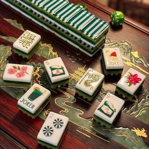 May include: A set of white and green Mahjong tiles featuring various illustrations including a dragon, flowers, and Joker cards with drink graphics. The tiles are arranged on a dark wood surface with gold inlay designs and a green die.