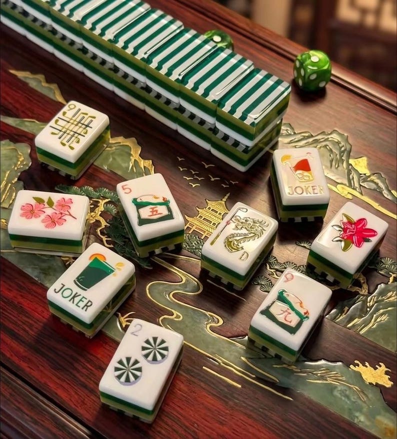 May include: A set of white and green Mahjong tiles featuring various illustrations including a dragon, flowers, and Joker cards with drink graphics. The tiles are arranged on a dark wood surface with gold inlay designs and a green die.