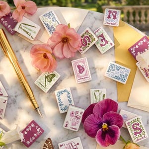K&ouml;nnte beinhalten: Eine Sammlung rechteckiger Mahjong-Steine mit verschiedenen Blumen-, Schmetterlings- und Buchstabenmotiven in rosa, violetter, gr&uuml;ner und blauer Tinte. Ein goldener F&uuml;llfederhalter liegt zwischen den Steinen, die zusammen mit rosa und violetten Blumen verstreut sind.