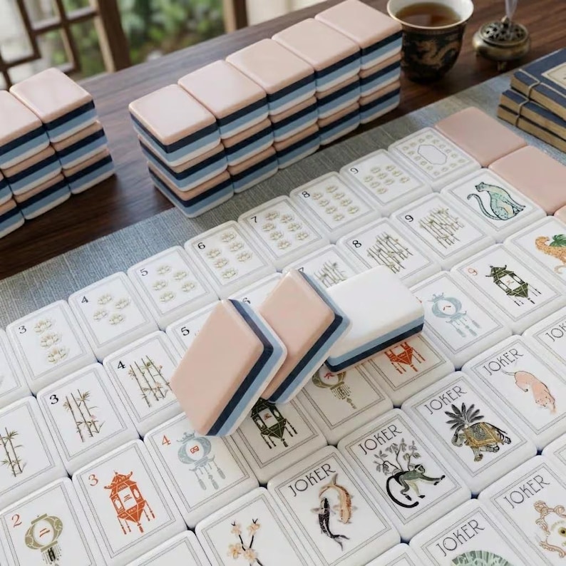 May include: A set of mahjong tiles featuring a multi-layered design with a pale pink top, a dark blue middle, and a white base. The tiles display various illustrations including bamboo, lanterns, and animals, with several tiles labeled as Joker.