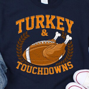 Op de afbeelding: Een marineblauw sweatshirt met ronde hals en een afbeelding van een gebraden kalkoen in de vorm van een American football, omringd door korenaren en de oranje tekst "TURKEY & TOUCHDOWNS". Het sweatshirt is gecombineerd met een lichtblauwe jeans en witte sneakers.