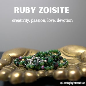 Könnte beinhalten: Eine grüne und rosafarbene Edelsteinkette mit einem silbernen Schiebeverschluss, die auf einer goldfarbenen, handförmigen Schale liegt. Die Kette besteht aus Rubin-Zoisit-Perlen und ist mit "Kreativität, Leidenschaft, Liebe, Hingabe" beschriftet.