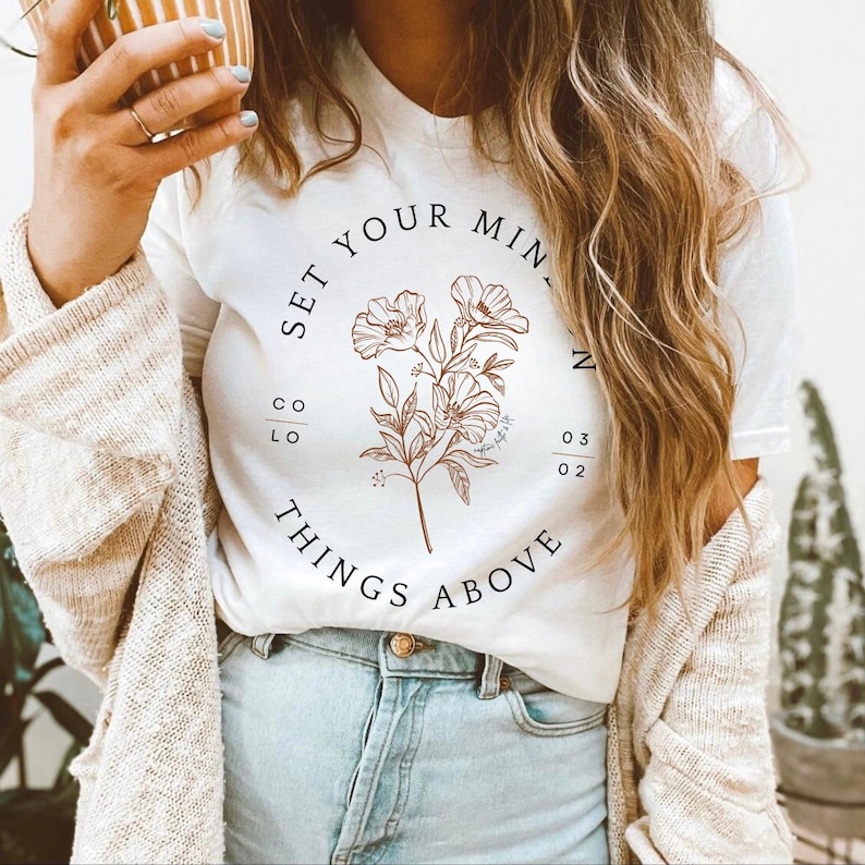 May include: White t-shirt with a floral design and the text "SET YOUR MIND ON THINGS ABOVE".