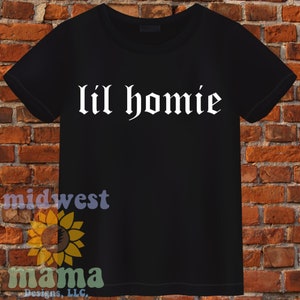 Peut inclure: T-shirt noir avec un texte blanc qui dit "lil homie" dans une police gothique.