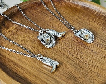 May include: Two silver necklaces with charms of a cowboy boot and a cowboy hat on a wooden surface. The necklaces are on a wooden surface with a green plant and white flowers in the background.