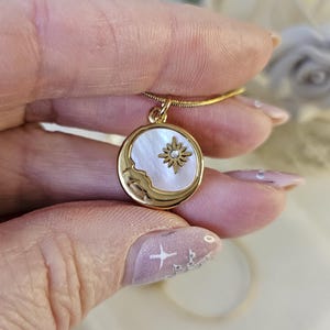 May include: A gold-coloured necklace with a circular pendant. The pendant features a mother-of-pearl moon and a sunburst design. The necklace is held by a hand, highlighting the jewellery's details. A delicate piece of jewellery.