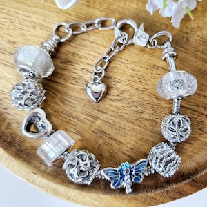 May include: A silver charm bracelet featuring a selection of charms, including a heart, a butterfly and a flower. The bracelet is displayed on a wooden surface.