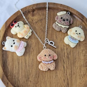 May include: A silver necklace with a pendant of a small, tan-coloured dog. The necklace is surrounded by four other dog-shaped charms in various colours, including white, tan, and brown. The charms are arranged on a wooden tray.