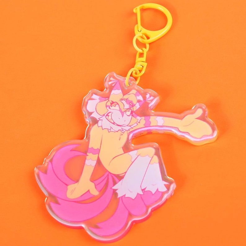 May include: A clear acrylic keychain featuring a cartoon character with yellow, pink, and white accents. The character has a fox-like tail and is attached to a yellow clasp.