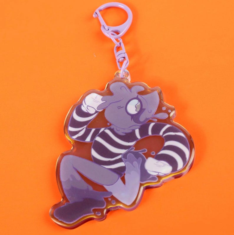 May include: A purple and white striped cartoon character keychain with a lavender clasp. The character has a blob-like body and is surrounded by a clear border. The background is a solid orange.