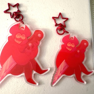 May include: Two red acrylic keychains featuring a cartoon character with a smiling face and a weapon. Each keychain has a red star-shaped clasp. The keychains are approximately 3 inches in length.