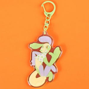 May include: A colorful keychain featuring a cartoon character with a green head and arms, a yellow body, and purple legs. The character is wearing a white shirt with green polka dots. The keychain has a green clasp and chain.