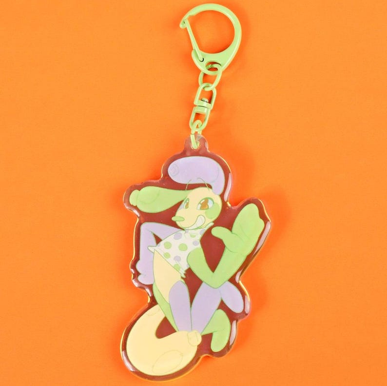 May include: A colorful keychain featuring a cartoon character with a green head and arms, a yellow body, and purple legs. The character is wearing a white shirt with green polka dots. The keychain has a green clasp and chain.
