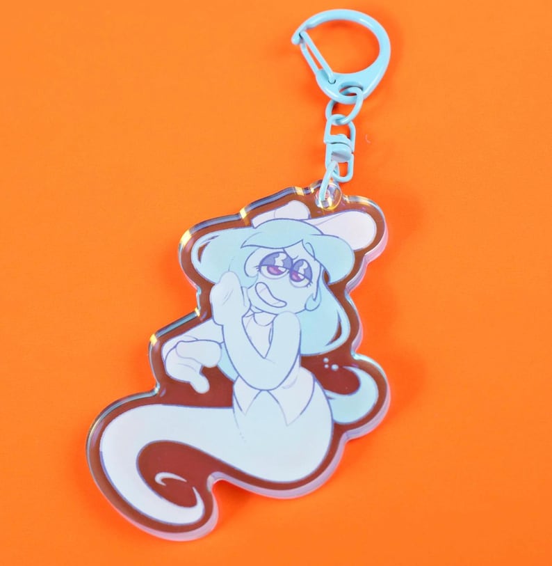 May include: A light blue keychain featuring a cartoon ghost character with a friendly expression. The ghost has light blue hair and a white outfit. The keychain has a blue clasp and chain, and is set against an orange background.