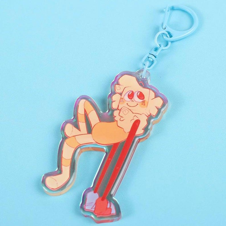 May include: A clear acrylic keychain featuring a cartoon character in shades of orange and red. The character is lying down and holding a red object. The keychain has a light blue clasp.