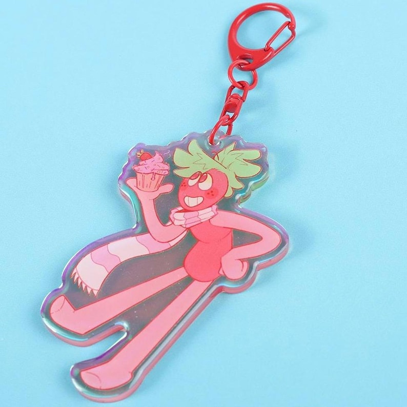 May include: A pink and red cartoon character keychain holding a cupcake. The character has green hair, a pink scarf, and a red keychain clasp. The keychain is made of clear acrylic with a holographic effect.