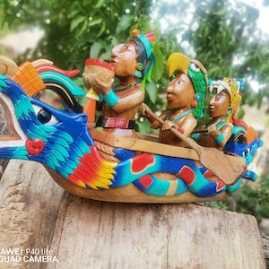 Könnte beinhalten: Eine Holzskulptur eines Bootes in Form eines Drachenkopfes mit drei Figuren im Inneren. Der Drache ist blau, rot und grün bemalt. Die Figuren tragen bunte Kopfbedeckungen und halten Paddel und eine Schale. Die Skulptur steht auf einer Holzoberfläche.