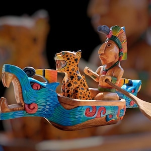 May include: A hand-carved wooden sculpture depicting a boat with a jaguar and a figure in traditional headdress. The boat's front is a blue serpent's head. The jaguar has orange and black spots. The figure is paddling with a wooden oar.
