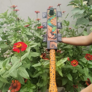 May include: A carved wooden ceremonial weapon with a detailed design. The top section features a green dragon carving and a human face, with a colorful serpent design. The handle is orange with black spots.