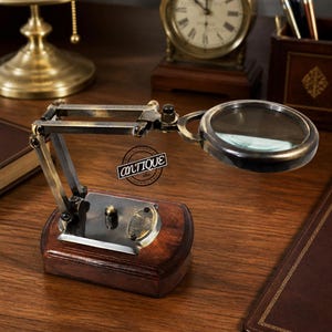 May include: A brass magnifying glass mounted on an adjustable articulated arm with a dark wooden base. The metal frame has a weathered finish, and a small oval plaque is attached to the base. The magnifying lens is circular and clear.