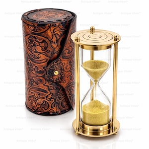 Könnte beinhalten: Eine Sanduhr aus Messing mit klaren Glaskolben, die feinen goldenen Sand enthalten, neben einem zylindrischen braunen Lederetui mit geprägtem Blumenmuster und einem einzelnen goldfarbenen Metalldruckknopf an der vorderen Klappe.