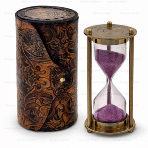 May include: A brass hourglass with purple sand positioned next to a cylindrical brown leather case featuring an embossed floral pattern and a small gold-coloured metal snap closure on the front side of the container.