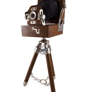 Puede incluir: Una cámara de madera vintage con un fuelle negro y detalles plateados, montada sobre un trípode. La cámara tiene un diseño clásico con una lente y varios diales. El trípode tiene patas de madera y una base de metal plateado, conectadas por una cadena.