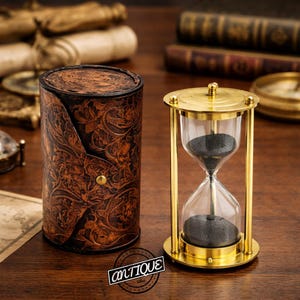 Könnte beinhalten: Eine Messing-Sanduhr mit schwarzem Sand neben einem zylindrischen braunen Lederetui mit geprägtem Blumenmuster. Das Etui hat einen einzelnen Metalldruckknopf. Der Text "antique" ist in einem kreisförmigen Logo unten zu sehen.