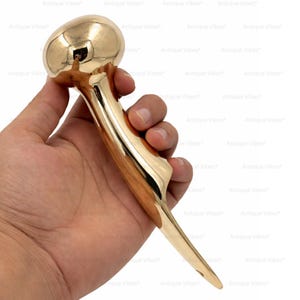 Könnte beinhalten: Ein polierter, goldfarbener Metall-Schuhlöffel mit einem glatten, geschwungenen Griff und einem abgerundeten, knollenförmigen Ende. Das Objekt hat eine glänzende, reflektierende Oberfläche und ein spitz zulaufendes Ende mit einem kleinen Loch.