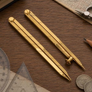May include: Two gold-toned metal drafting compasses with pointed tips and adjustable screw mechanisms. The instruments feature a sleek, elongated design with a smooth finish, positioned diagonally across the frame alongside drafting tools.