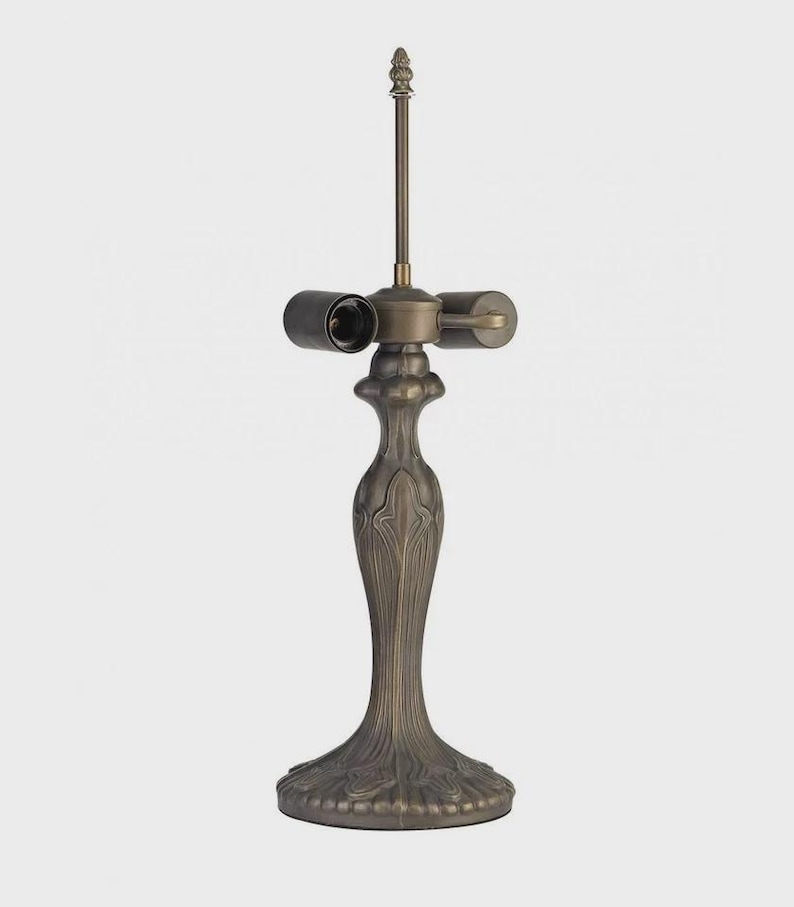 May include: A bronze-colored lamp base with a decorative, Art Nouveau-inspired design. The base features a detailed, textured pattern and a slender, elongated stem. The lamp has two bulb sockets and a central rod topped with a decorative finial.